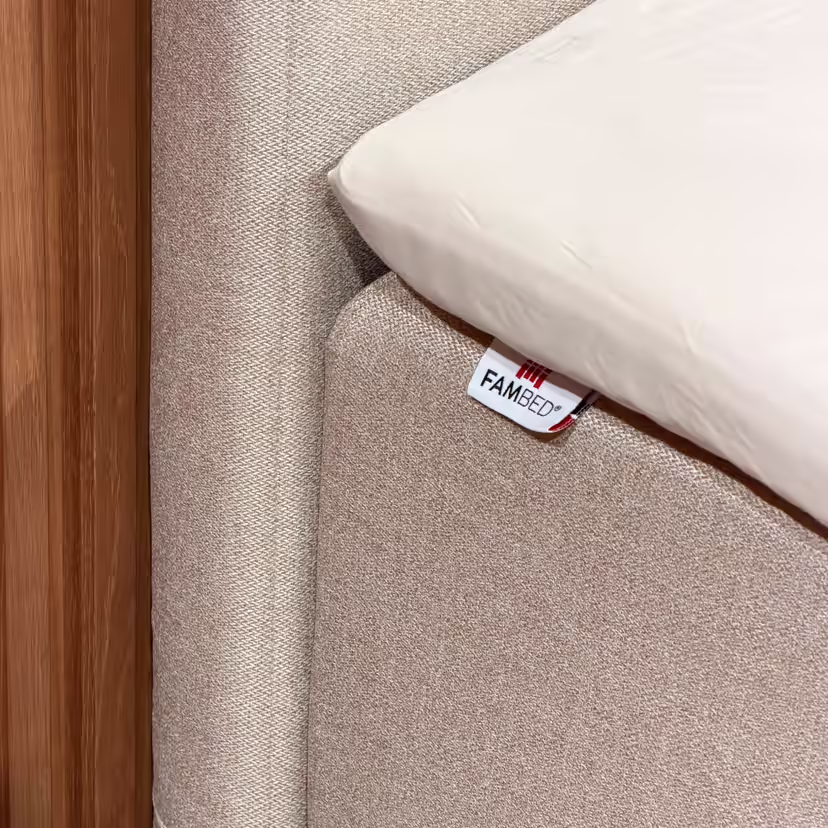 Nærbillede af det beige cover om sengens hjørne, med FAMBED-mærke og hvidt lagen ovenpå